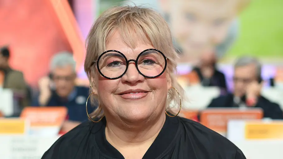 Ilka Bessin Eine lächelnde Frau mit kurzen blonden Haaren, runder schwarzer Brille und großen Creolen trägt eine schwarze Jacke.