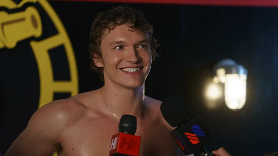 Ilya steht oberkörperfrei vor einem schwarzen Hintergrund mit gelbem Logo. Er wird von mehreren Mikrofonen interviewt und wirkt entspannt und charmant.