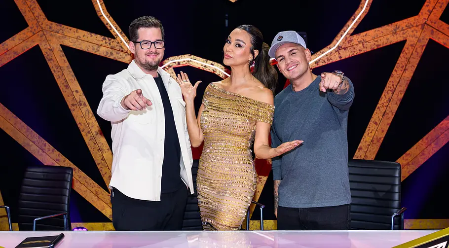 Im Rateteam machte sich Pietro Lombardi zumindest ziemlich gut. Chris Tall, Verona Pooth und Pietro Lombardi posieren hinter einem Pult vor einer orangefarbenen, leuchtenden Maskenkulisse.