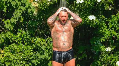 Nico Legat steht mit tätowiertem Oberkörper und schwarzer Unterhose unter einer Dusche im Grünen und hält die Hände auf den Kopf. - Foto: RTL