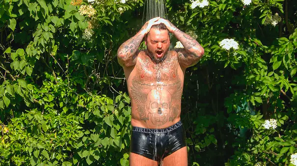 Nico Legat steht mit tätowiertem Oberkörper und schwarzer Unterhose unter einer Dusche im Grünen und hält die Hände auf den Kopf. - Foto: RTL