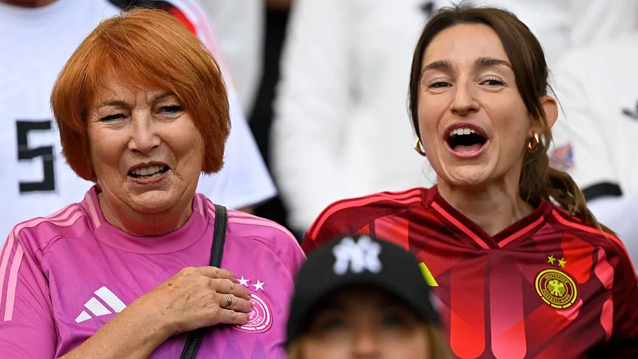 Burgi Nagelsmann Zwei Fans der deutschen Nationalmannschaft stehen auf der Tribüne eines Fußballstadions und jubeln begeistert. Die ältere Frau trägt ein pinkes DFB-Trikot, die jüngere Frau ein rot-schwarzes Deutschland-Trikot. Beide verfolgen das Spielgeschehen aufmerksam und wirken emotional mitgerissen.