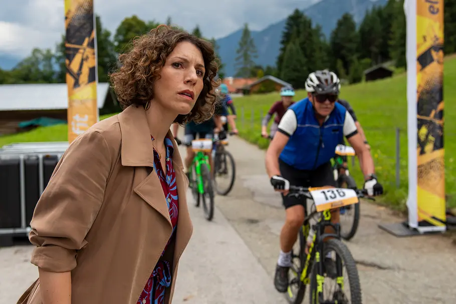 Einfach Elli Einfach Elli - Verzeihen: Vor einer Berglandschaft: Karin Schuster (Eva-Maria Reichert) steht am Ziel des Radrennens und hält Ausschau nach ihrem Freund. Einige Fahrer (Komparsen) des Radrennens fahren neben ihr durch das Ziel.