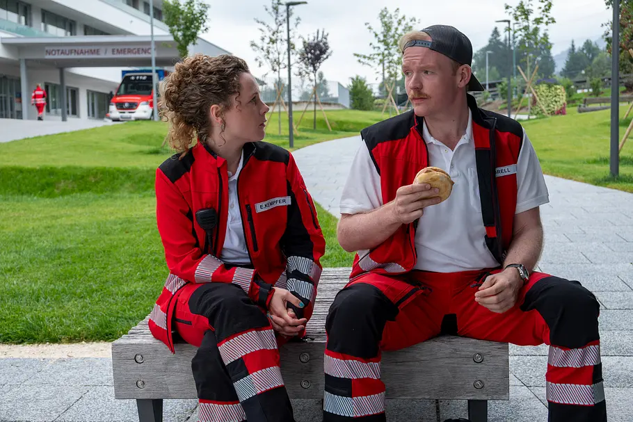 Einfach Elli Einfach Elli - Neuanfang: Elli Kempfer (Klara Deutschmann) und Felix Sorell (Sebastian Griegel) machen nach einem Rettungseinsatz eine Pause. Sie sitzen vor der Klinik in einer Grünanlage auf einer Bank und schauen sich an. Sebastian Griegel hat ein belegtes Brötchen in seiner rechten Hand.