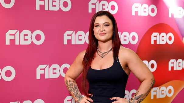 Sophia Thiel posiert mit den Händen an den Hüften im schwarzen Top vor einer pinken FIBO-Wand. Ihre Arme sind tätowiert. - Foto: Imago/Panama Pictures Sophia Thiel posiert mit den Händen an den Hüften im schwarzen Top vor einer pinken FIBO-Wand. Ihre Arme sind tätowiert. - Foto: Imago/Panama Pictures