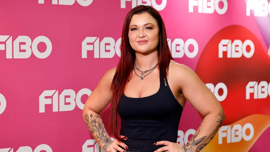 Sophia Thiel versteckt ihre Probleme nicht vor ihren Followern. Sophia Thiel posiert mit den Händen an den Hüften im schwarzen Top vor einer pinken FIBO-Wand. Ihre Arme sind tätowiert.