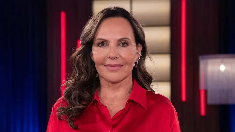 Natascha Ochsenknecht Natascha Ochsenknecht trägt eine rote Bluse und langes, gewelltes Haar. Hinter ihr leuchten rote und blaue Lichter.