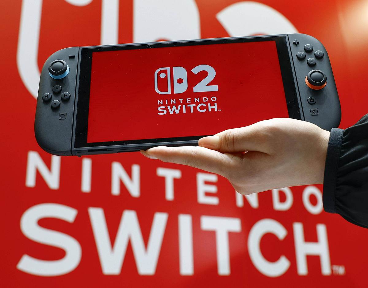 Ein Foto eines Nintendo-Switch 2-Handhelds mit dem Switch 2-Logo auf dem Display, gehalten von einer Hand vor einem Nintendo Switch 2 Plakat im Hintergrund