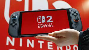 Ein Foto eines Nintendo-Switch 2-Handhelds mit dem Switch 2-Logo auf dem Display, gehalten von einer Hand vor einem Nintendo Switch 2 Plakat im Hintergrund - Foto:  IMAGO / Kyodo News