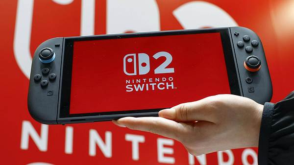 Ein Foto eines Nintendo-Switch 2-Handhelds mit dem Switch 2-Logo auf dem Display, gehalten von einer Hand vor einem Nintendo Switch 2 Plakat im Hintergrund - Foto:  IMAGO / Kyodo News