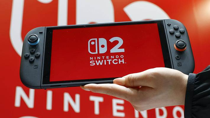 Ein Foto eines Nintendo-Switch 2-Handhelds mit dem Switch 2-Logo auf dem Display, gehalten von einer Hand vor einem Nintendo Switch 2 Plakat im Hintergrund - Foto:  IMAGO / Kyodo News