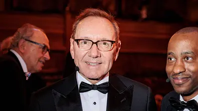 Kevin Spacey trägt einen schwarzen Anzug mit schwarzer Fliege. Neben ihm stehen zwei Männer. Er lächelt in die Kamera. - Foto:  IMAGO / ANP