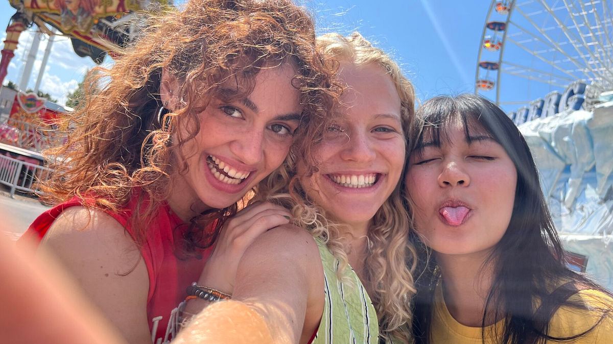 Inken, Vicky und Lena stehen auf einem Jahrmarkt und machen ein Selfie, grinsen dabei in die Kamera, Lena macht eine freche Grimasse.  