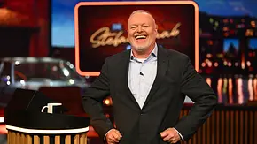Stefan Raab steht lachend im dunklen Anzug mit den Händen an den Hüften neben einem Pult im bunt beleuchteten Studio. - Foto: RTL / Raab Entertainment / Willi Weber Stefan Raab steht lachend im dunklen Anzug mit den Händen an den Hüften neben einem Pult im bunt beleuchteten Studio. - Foto: RTL / Raab Entertainment / Willi Weber