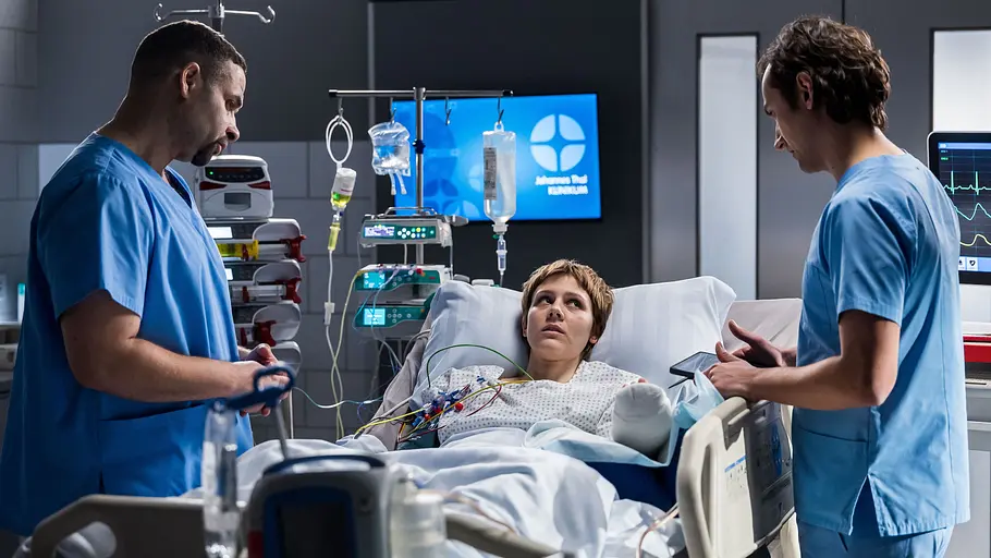 Zwei Männer in blauer Klinikkleidung stehen am Bett einer Patientin mit bandagiertem Arm. Daneben sind Infusionen und Monitore.