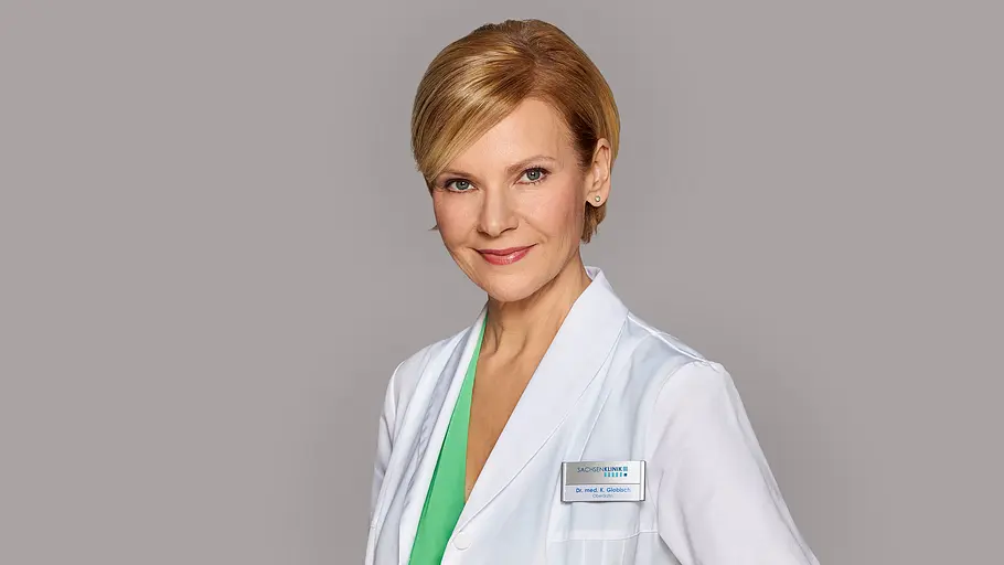 Andrea Kathrin Loewig trägt einen weißen Arztkittel über einem grünen Oberteil und lächelt vor grauem Hintergrund.