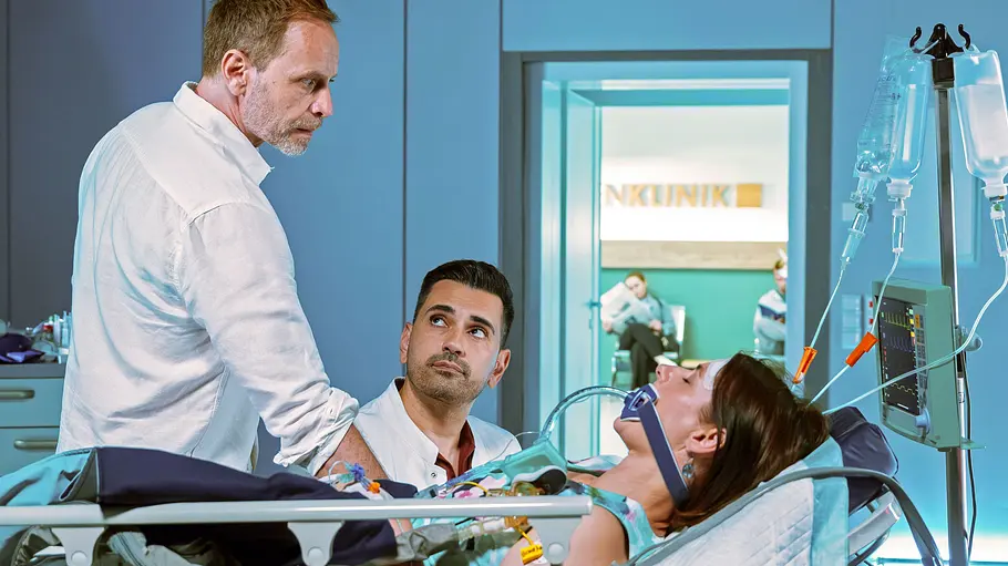 Kai schaut Maria verzweifelt an, die im Krankenbett liegt. Im Hintrrgrund ist Illay. 