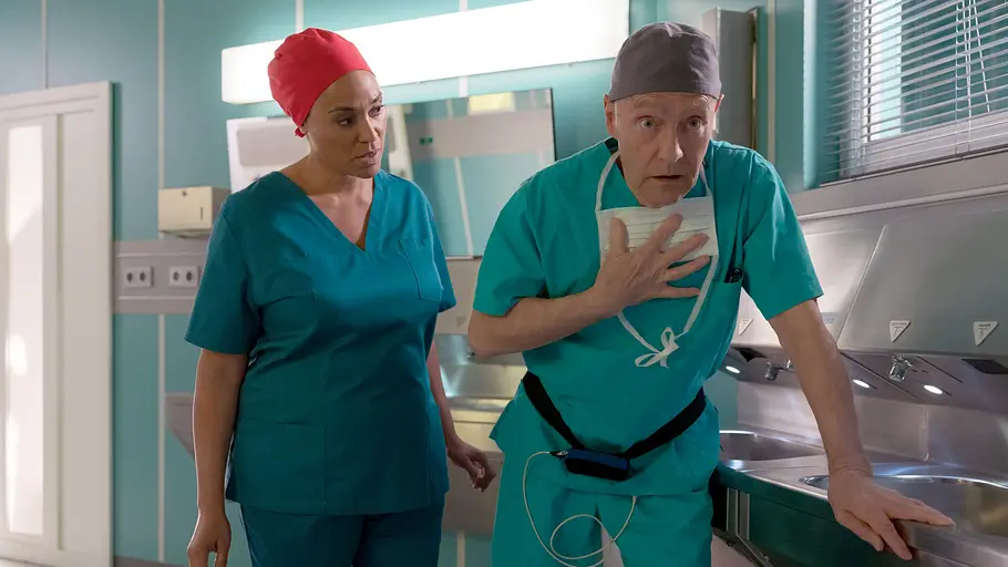 In aller Freundschaft Vorschau 27.10. - 1.12. Dr. Lucia Böhm sieht zu Dr. Rolf Kaminski, der in OP-Kleidung eine Hand an die Brust hält und sich am Waschbecken abstützt.