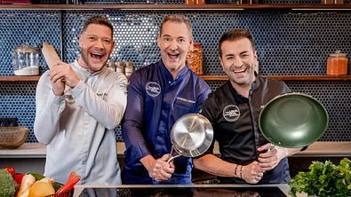 Robin Pietsch, Christian Henze und Ali Güngörmüs lachen in Kochjacken und halten einen Holzlöffel, einen Topf und eine Pfanne. - Foto: Joyn/Nadine Rupp Robin Pietsch, Christian Henze und Ali Güngörmüs lachen in Kochjacken und halten einen Holzlöffel, einen Topf und eine Pfanne. - Foto: Joyn/Nadine Rupp