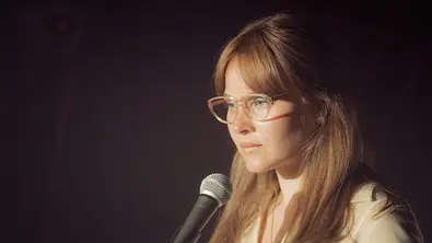 Kudamm 77 - Meine unbekannte Familie: Monika Franck-Schöllack (Sonja Gerhardt) steht vor dunklem Hintergrund an einem Mikrofon. Sie sieht unsicher nach vorne. - Foto: ZDF/Conny Klein