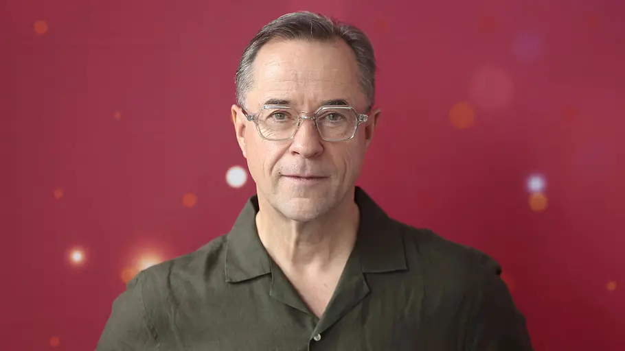 In seinem Podcast „Liefers + Broicher“ kann sich Jan Josef Liefers zu allen aktuellen Themen äußern. Jan Josef Liefers trägt eine helle Brille und ein dunkelgrünes Hemd vor einem roten Hintergrund mit unscharfen Lichtpunkten.