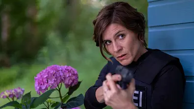 In Wahrheit - Die Liebe und der Tod: Judith Mohn (Christina Hecke) steht dicht an einer Holzhütte und zielt mit ihrer Dienstwaffe nach vorne. Neben ihr blühen auffällige rosafarbene Hortensien, während der Hintergrund von dichtem Grün umgeben ist. Die Szene wirkt angespannt und konzentriert. - Foto: ZDF/Manju Sawhney