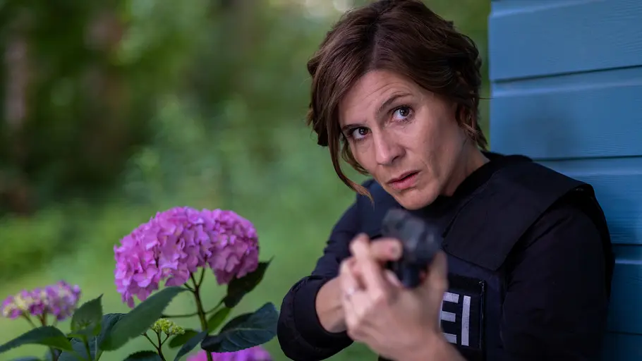 In Wahrheit - Die Liebe und der Tod: Judith Mohn (Christina Hecke) In Wahrheit - Die Liebe und der Tod: Judith Mohn (Christina Hecke) steht dicht an einer Holzhütte und zielt mit ihrer Dienstwaffe nach vorne. Neben ihr blühen auffällige rosafarbene Hortensien, während der Hintergrund von dichtem Grün umgeben ist. Die Szene wirkt angespannt und konzentriert.