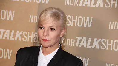 Ina Müller trägt eine schwarze Jacke, eine weiße Bluse und große Creolen vor einer Wand mit „NDR Talk Show“-Schriftzügen. - Foto: IMAGO / Andre Lenthe