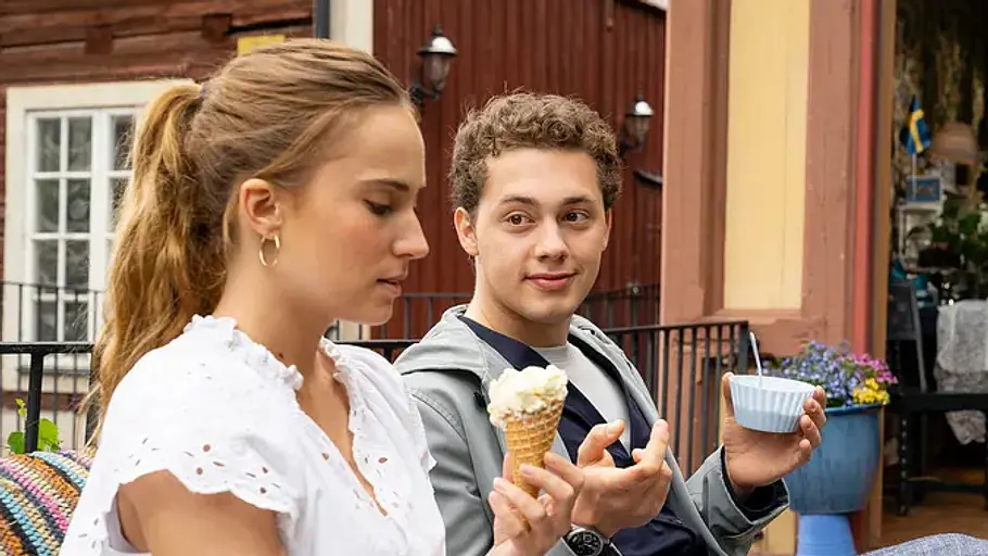 Sommerflirt bei „Inga Lindström: Klang der Sehnsucht“ Zwei junge Menschen sitzen draußen vor einem Café, essen Eis und unterhalten sich in einer Szene aus „Inga Lindström: Klang der Sehnsucht“.