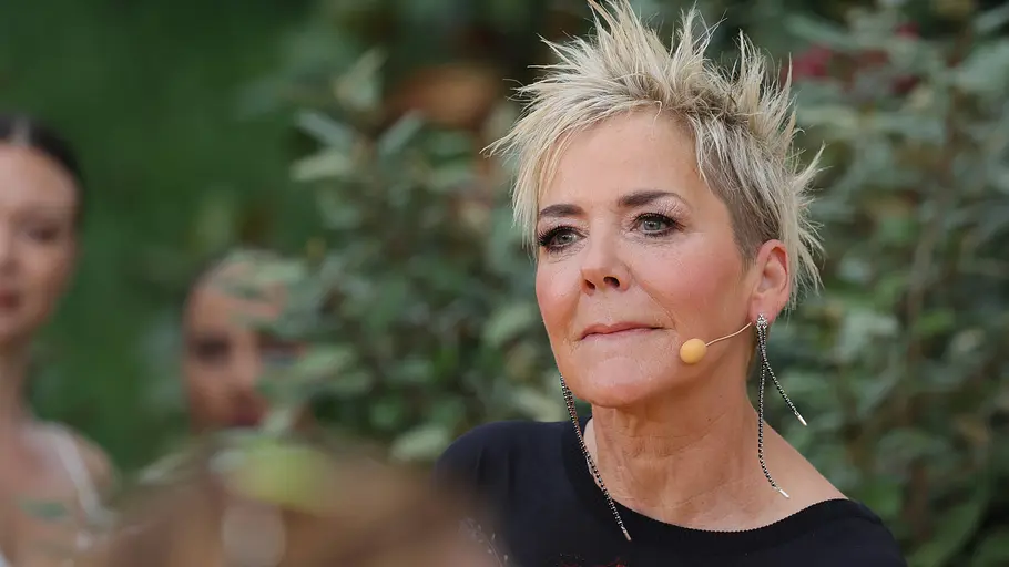 Inka Bause: Warum sie selbst keinen Mann kennenlernt Inka Bause trägt blonde, kurze Haare, ein Headset und ein schwarzes Oberteil mit Blumenmotiv. Hinter ihr stehen weitere Personen.