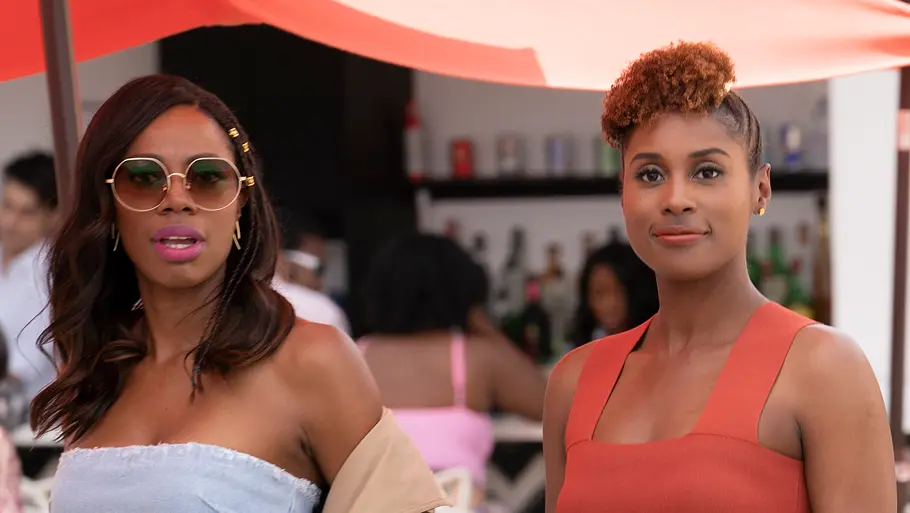 Insecure Zwei stilvoll gekleidete Frauen stehen in einem Beachclub vor einer Bar und blicken aufmerksam nach vorne. Eine trägt ein hellblaues Jeans-Outfit mit gemusterter Jacke und Sonnenbrille, die andere ein orangefarbenes Crop-Top mit geblümtem Rock.