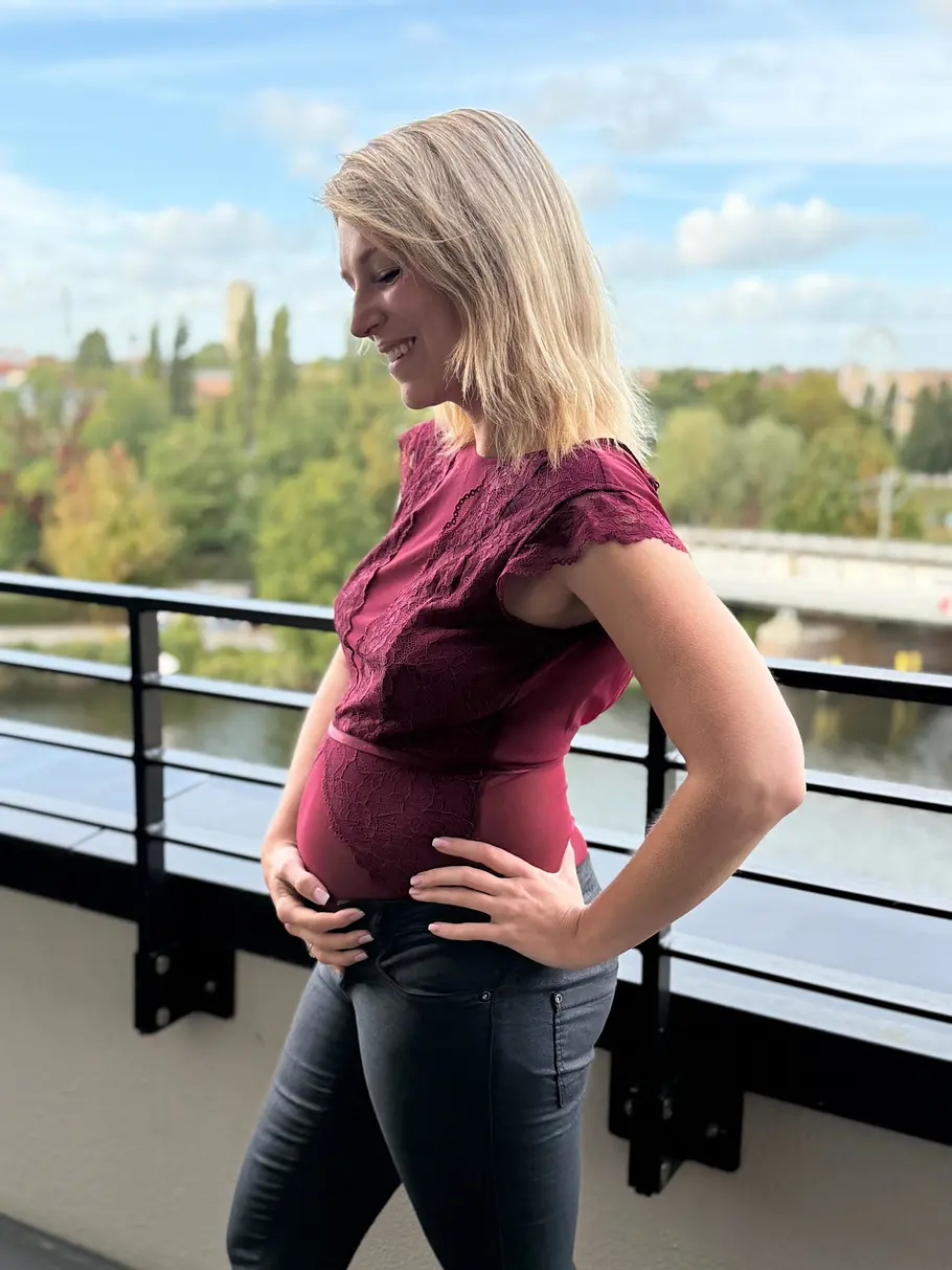 iris-mareike-steen-schwanger Iris Mareike Steen steht lächelnd im weinroten Spitzenoberteil auf einem Balkon, die Hände an Bauch und Hüfte gelegt.