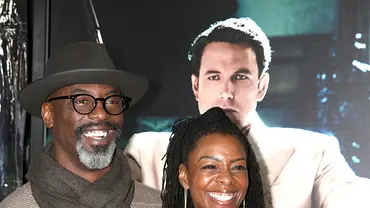 Isaiah Washington mit Hut, Brille und Schal und seine Frau mit langen Haaren lächeln vor einem Plakat mit einem Mann im hellen Anzug. - Foto:  IMAGO / Independent Photo Agency