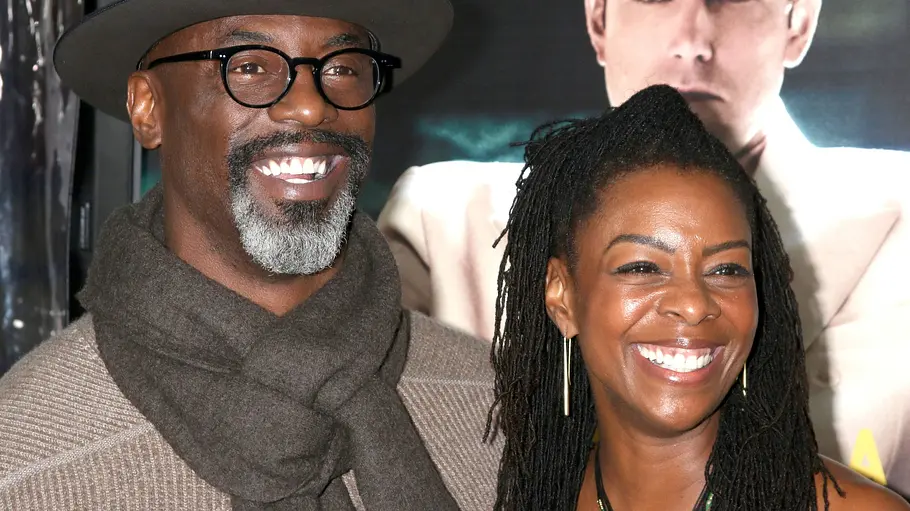 Isaiah Washington und seine Frau waren 29 Jahre verheiratet. Isaiah Washington mit Hut, Brille und Schal und seine Frau mit langen Haaren lächeln vor einem Plakat mit einem Mann im hellen Anzug.