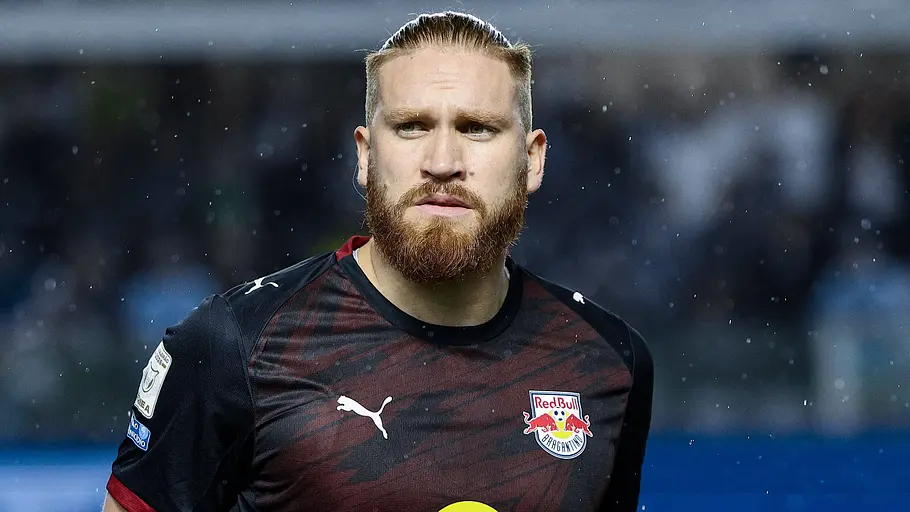 Fußballspieler von Red Bull Salzburg im schwarz-roten Trikot mit Red-Bull-Logo blickt während eines Spiels konzentriert nach vorne. Im Hintergrund sind der Rasen, leichte Regentropfen und die verschwommenen Zuschauerränge zu sehen.