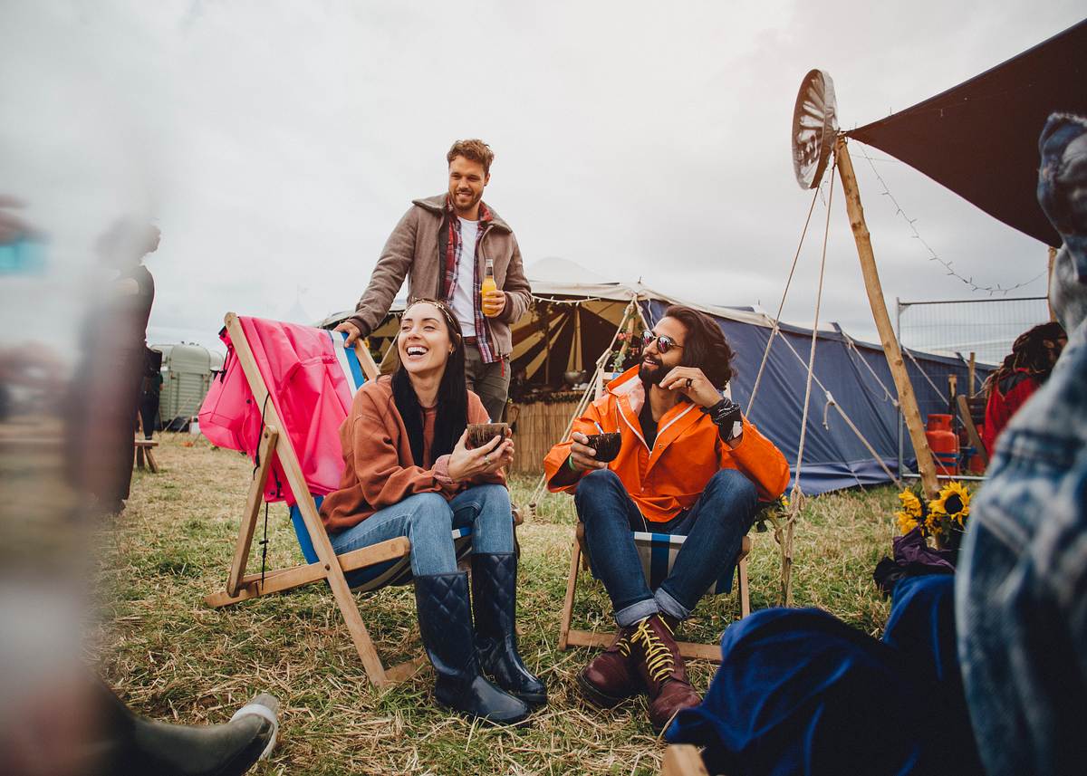 Der Festival-Sommer steht vor der Tür. Schon die passende Ausrüstung parat? Drei Menschen sitzen auf einem Festival-Zeltplatz beieinander, zwei davon in Campingstühlen.
