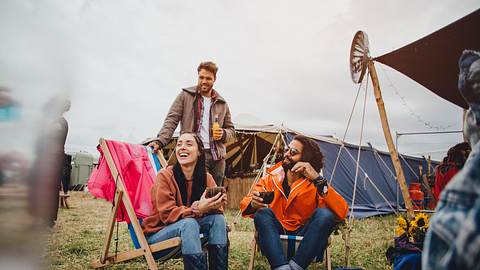 Drei Menschen sitzen auf einem Festival-Zeltplatz beieinander, zwei davon in Campingstühlen. - Foto: iStock / SolStock