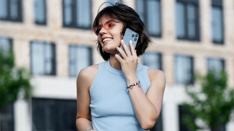Eine junge Frau mit hellblauen Oberteil und einer roten Sonnenbrille hält sich lächelnd ein iPhone an das Ohr. - Foto: iStock / YakubovAlim