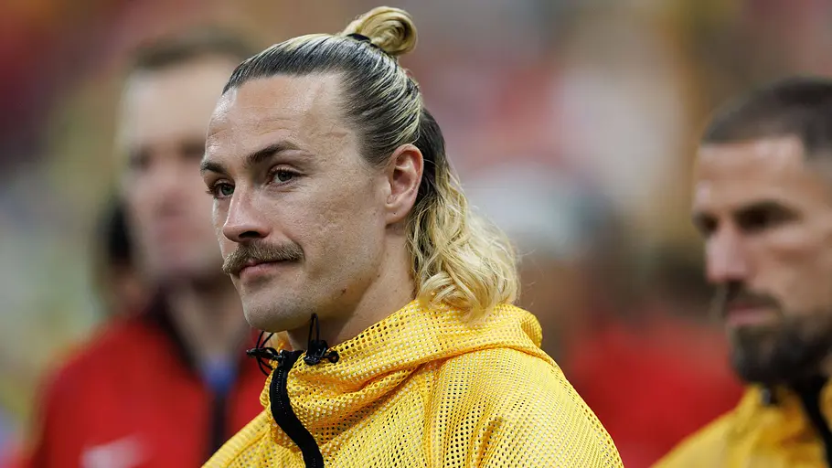 Ein australischer Nationalspieler mit blondem, zum Dutt gebundenem Haar und Schnurrbart trägt vor dem Spiel eine gelbe Trainingsjacke mit Verbandslogo. Im Hintergrund sind weitere Teammitglieder unscharf zu sehen.