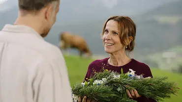Die Maiwald: Beziehungsweise: Auf einer Almwiese: Strahlend steht Dr. Johanna Maiwald (Doris Schretzmayer) Jakob Reindl (Bernhard Piesk) gegenüber. Dieser steht mit dem Rücken zur Kamera. Sie hält ein Gesteck aus Fichtenzweigen und Blumen in den Händen und lächelt Jakob glücklich an. Im Hintergrund grast eine Kuh. - Foto: ZDF/Lukas-Frederik Pfaff Die Maiwald: Beziehungsweise: Auf einer Almwiese: Strahlend steht Dr. Johanna Maiwald (Doris Schretzmayer) Jakob Reindl (Bernhard Piesk) gegenüber. Dieser steht mit dem Rücken zur Kamera. Sie hält ein Gesteck aus Fichtenzweigen und Blumen in den Händen und lächelt Jakob glücklich an. Im Hintergrund grast eine Kuh. - Foto: ZDF/Lukas-Frederik Pfaff