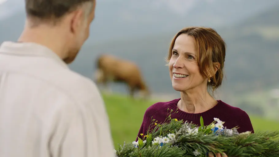 Jakob Reindl (Bernhard Piesk) und Dr. Johanna Maiwald (Doris Schretzmayer) bei „Die Maiwald“. Die Maiwald: Beziehungsweise: Auf einer Almwiese: Strahlend steht Dr. Johanna Maiwald (Doris Schretzmayer) Jakob Reindl (Bernhard Piesk) gegenüber. Dieser steht mit dem Rücken zur Kamera. Sie hält ein Gesteck aus Fichtenzweigen und Blumen in den Händen und lächelt Jakob glücklich an. Im Hintergrund grast eine Kuh.