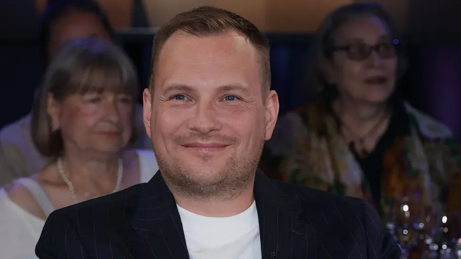 Jakob Schwerdtfeger Jakob Schwerdtfeger sitzt entspannt in einem Sessel im Studio einer Talkshow und lächelt in die Kamera. Er trägt einen dunklen Nadelstreifenanzug mit weißem T‑Shirt. Im Hintergrund sind unscharf weitere Gäste und gedeckte Tische mit Gläsern zu sehen.