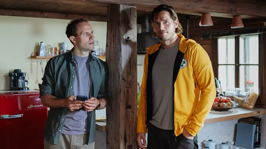 Jan Fugain Arne Löber Bergretter Jan hält links ein Smartphone, Markus steht rechts in gelber Jacke. Zwischen ihnen steht ein Holzbalken in einer Küche.