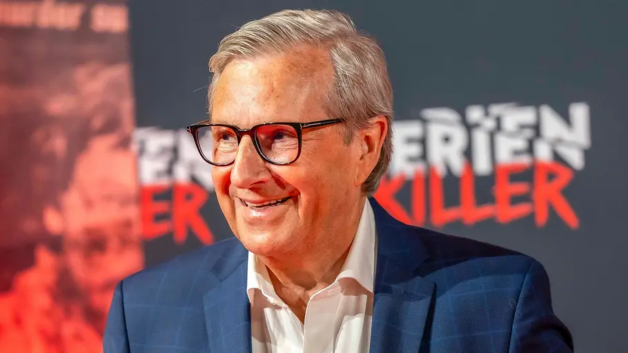 Jan Hofer HSE Jan Hofer mit Brille lächelt im blau karierten Sakko und weißen Hemd vor einer Wand mit rot-weißer Schrift.