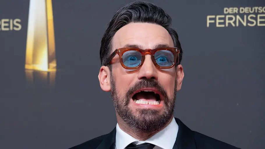 Jan Köppen Quietscheenten-Wette. Jan Köppen mit blau getönter Brille und schwarzem Anzug posiert mit offenem Mund vor einer Wand des Deutschen Fernsehpreises.