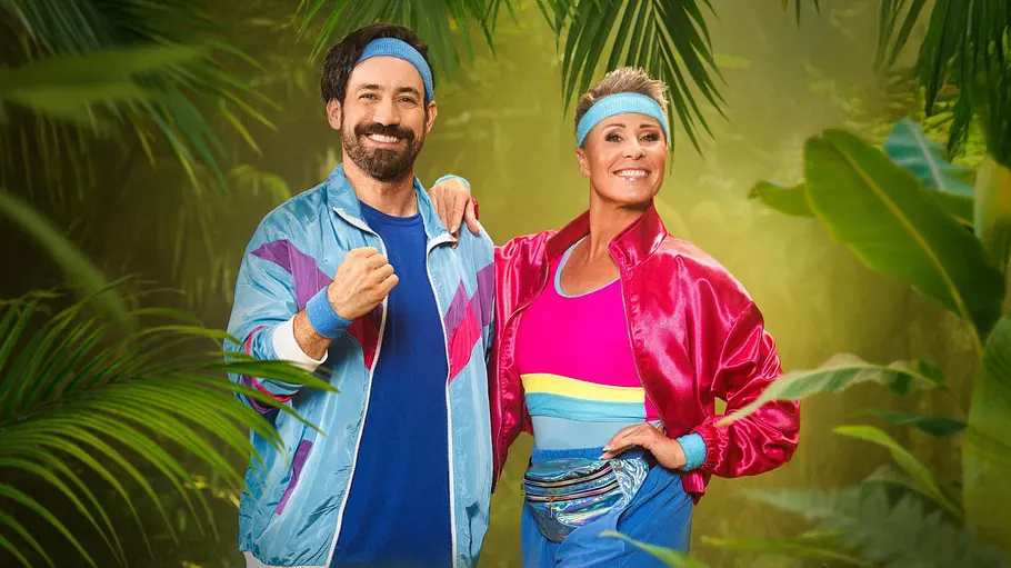 Jan Köppen und Sonja Zietlow moderieren das „Dschungelcamp“. Jan Köppen und Sonja Zietlow posieren lächelnd in bunten Sportoutfits mit blauen Stirnbändern vor tropischen Pflanzen.