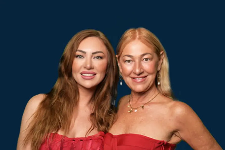 Jana Maria und Eva-Maria Herz stehen in roten, schulterfreien Kleidern vor dunkelblauem Hintergrund und lächeln in die Kamera.