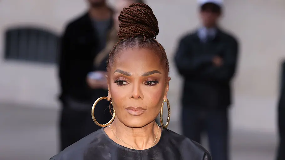 Janet Jackson trägt einen hohen Dutt aus geflochtenen Zöpfen, große goldene Creolen und ein schwarzes, glänzendes Oberteil.
