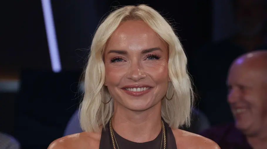 Janin Ullmann Janin Ullmann sitzt im Studio einer Talkshow und lächelt in die Kamera. Sie trägt ein ärmelloses braunes Outfit mit goldenen Ketten und mehreren Ringen; im Vordergrund stehen Wassergläser und ein Blumenstrauß, im Hintergrund ist das Publikum unscharf zu sehen.