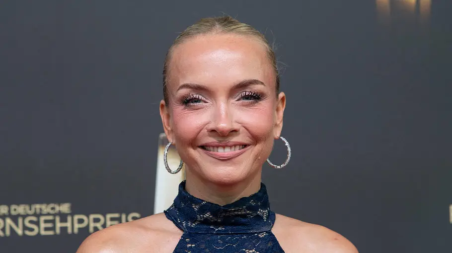 Janin Ullmann lächelt in einem dunkelblauen Spitzenkleid mit großen Creolen vor einer Wand mit dem Schriftzug „Deutscher Fernsehpreis“.